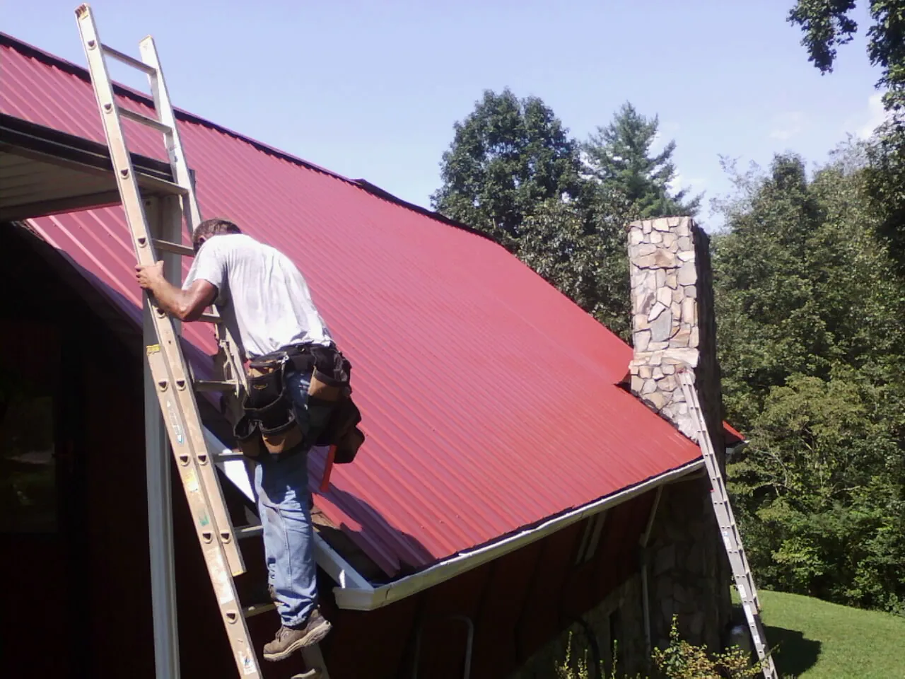 Roofer on a ladder beside a red metal roof and stone chimney — Soffit & Fascia Repair by Roofing Brothers in Bethany Beach